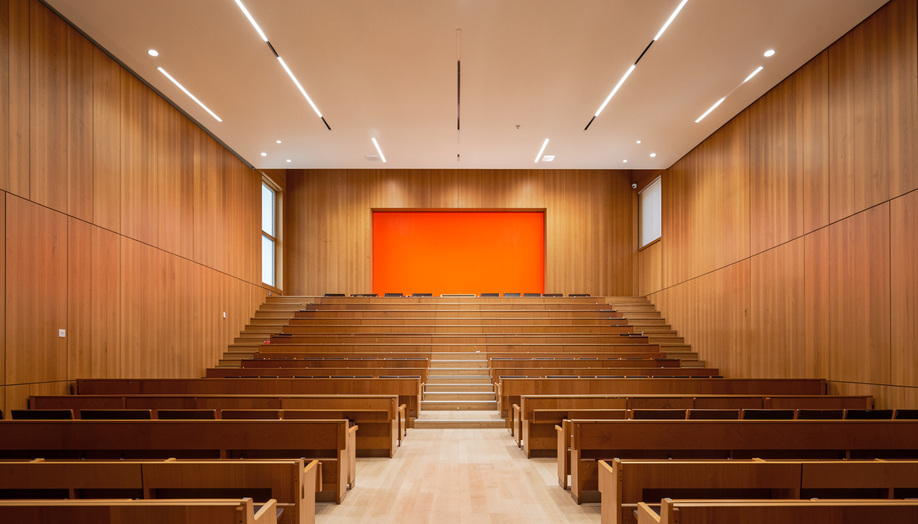 A clean architectural photograph with strong symmetry: a modern university lecture hall interior with tiered seating, warm wood tones, and soft overhead lighting